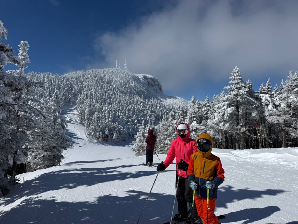 Two skiers in bright winter jackets (an adult in pink and a child in orange-yellow) stand on a sunlit groomed ski run surrounded by snow-covered evergreen trees, with a forested mountain peak topped by antennas and a blue sky and clouds overhead.