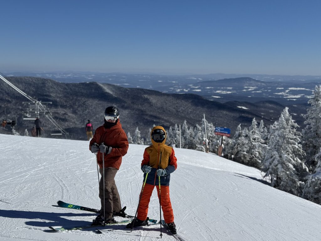 Two skiers—a taller adult in a rust jacket and brown pants and a child in a yellow-and-orange jacket—stand on a groomed snowy slope wearing helmets and goggles, with a chairlift to the left, snow-covered trees and a trail sign to the right, and wide snow-dusted mountains beneath a clear blue sky in the background.