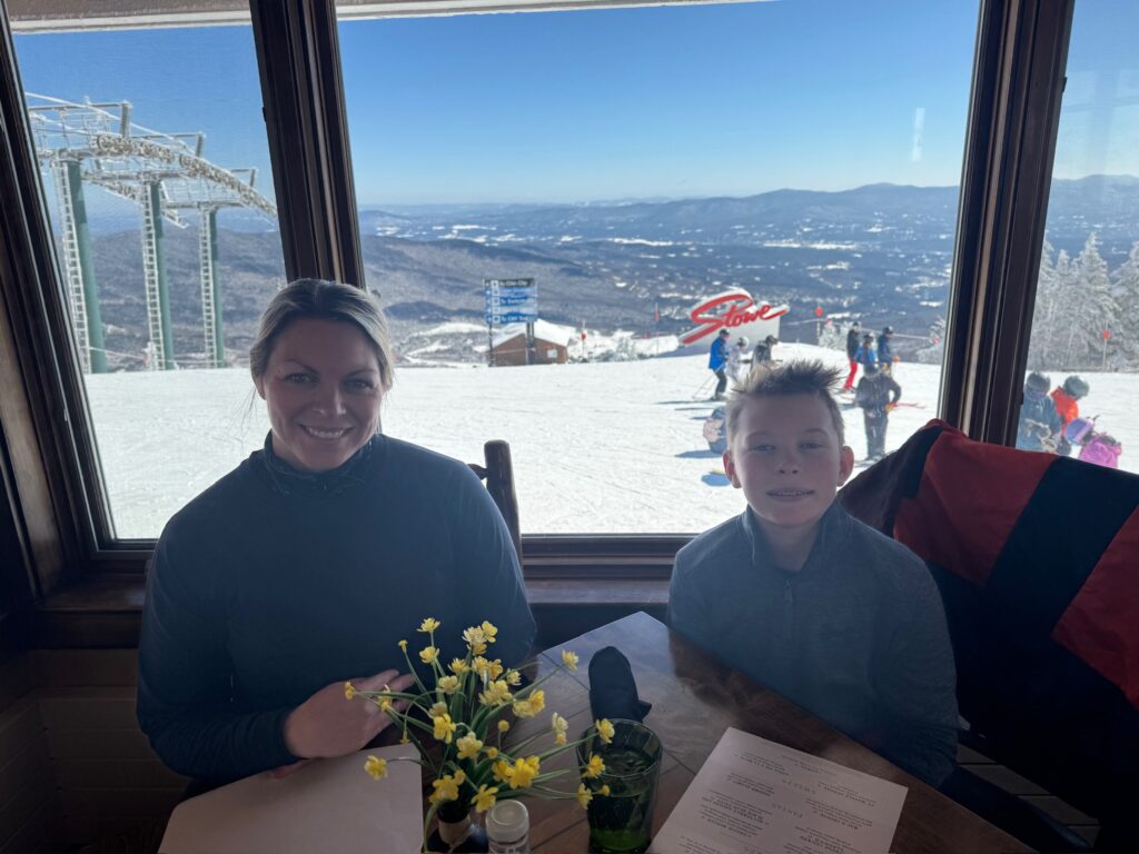 Woman and young boy seated at a wooden table in a mountaintop ski lodge with yellow flowers, menus and a green glass on the table, a large window behind them revealing a snowy slope with skiers, a red “Stowe” sign, ski-lift towers and a distant mountain range.