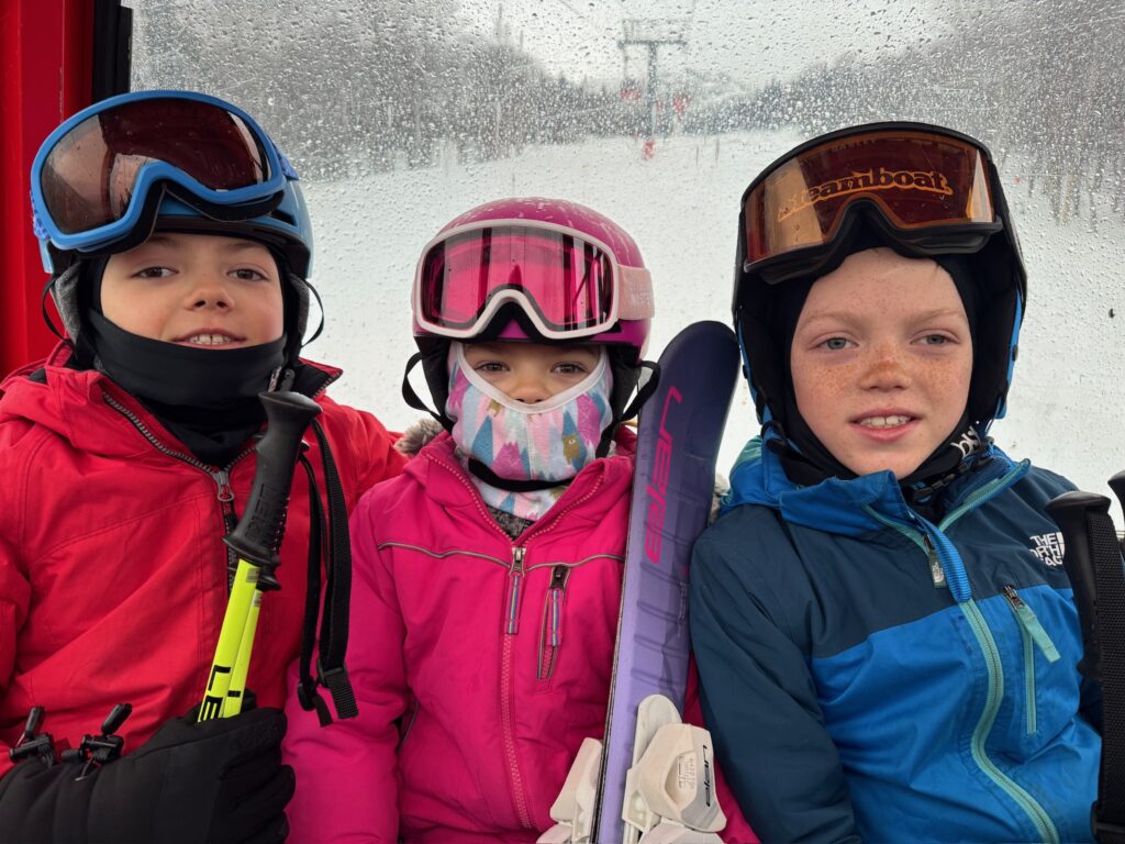Three children wearing ski helmets, goggles, and colorful winter jackets sit close together inside a gondola holding skis and poles, with a snow-speckled window and snowy slope visible behind them.