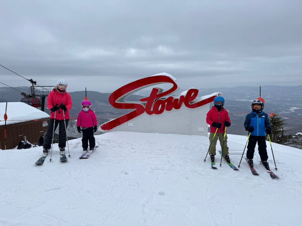 Four skiers in colorful jackets, helmets and goggles stand on a snowy summit holding poles in front of a large red “Stowe” sign, with an overcast sky, distant mountain ridges and a gondola lift visible to the left.