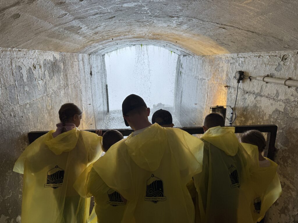 A group of people seen from behind wearing bright yellow rain ponchos with a black tour logo, leaning on a railing inside a damp concrete tunnel and looking out toward a bright curtain of falling water.