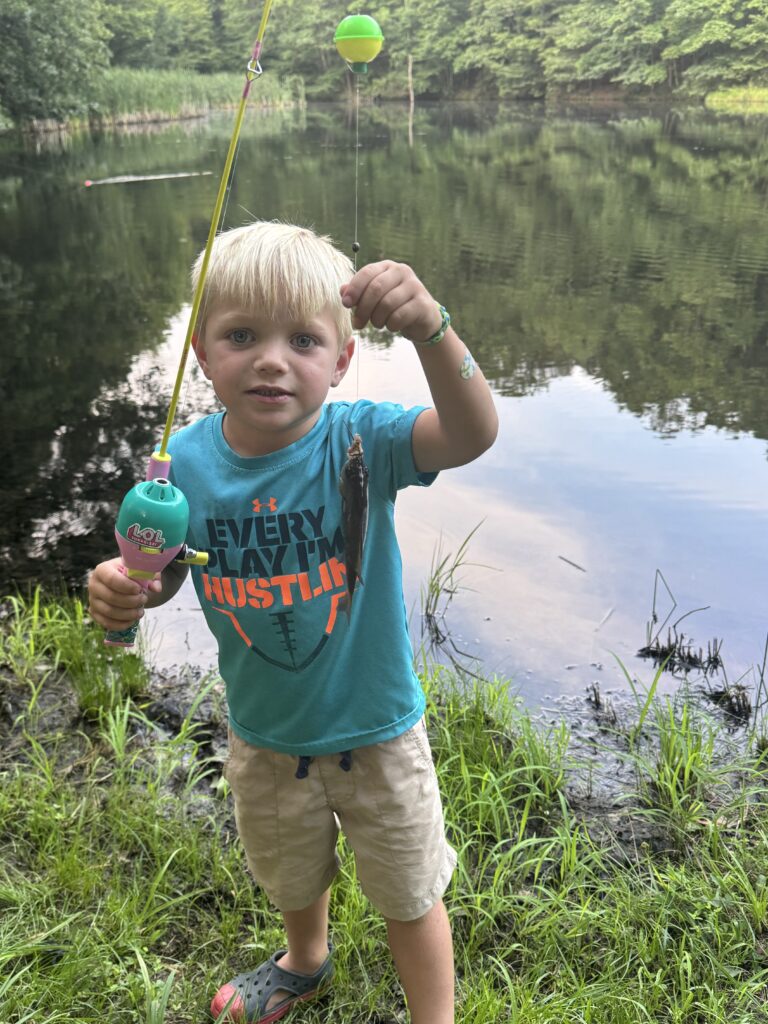 Young blonde boy standing on a grassy pond bank, holding a bright toy fishing rod in one hand and a small fish dangling from the line with the calm tree-lined pond reflected behind him.