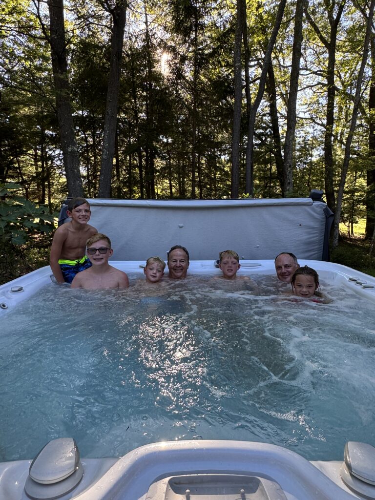A group of children and two adult men relax and smile in a bubbling outdoor hot tub set on the edge of a sunlit forest with tall trees and dappled light behind them.