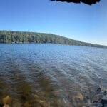 Panoramic view of a sunlit lake with rippling blue water, a tree-covered hill across the shore, a rocky shoreline in the foreground, and the bright sun shining through trees on the right casting long shadows on the rocks.
