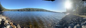 Panoramic view of a sunlit lake with rippling blue water, a tree-covered hill across the shore, a rocky shoreline in the foreground, and the bright sun shining through trees on the right casting long shadows on the rocks.
