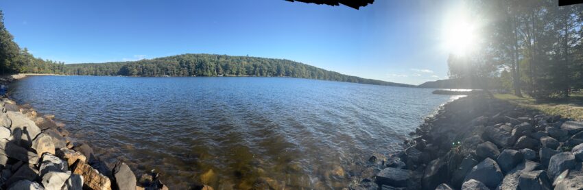 Panoramic view of a sunlit lake with rippling blue water, a tree-covered hill across the shore, a rocky shoreline in the foreground, and the bright sun shining through trees on the right casting long shadows on the rocks.