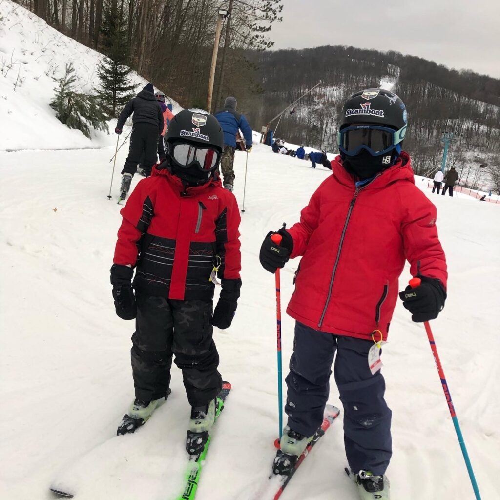 Two children in red ski jackets and black helmets with goggles standing on skis and holding poles on a snowy slope, with other skiers, trees and snow-covered hills in the background.