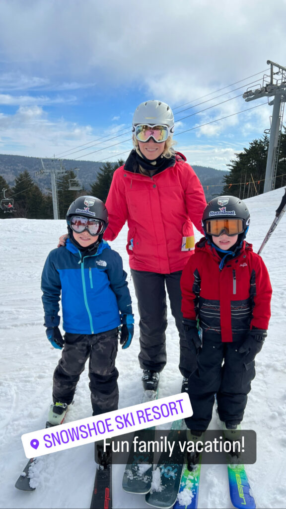 Woman in a white ski helmet and reflective goggles wearing a red jacket stands between two children in helmets and winter jackets on a snowy slope with their skis, chairlift cables and forested hills visible under a partly cloudy sky.