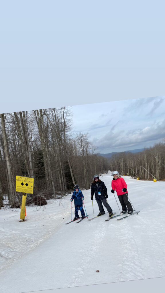 Three skiers — a child in blue, an adult in a dark jacket, and an adult in a bright pink jacket — stand on a groomed snowy ski trail beside a yellow caution sign reading “Shay’s Revenge,” with bare trees and distant hills under a cloudy sky.