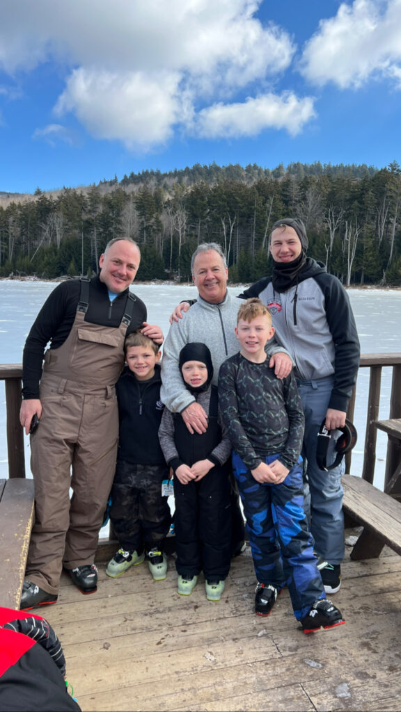 Three adult men and three boys in winter clothing and ski boots stand smiling with arms around each other on a wooden deck by a frozen lake, with a forested hillside and blue sky with clouds behind them.