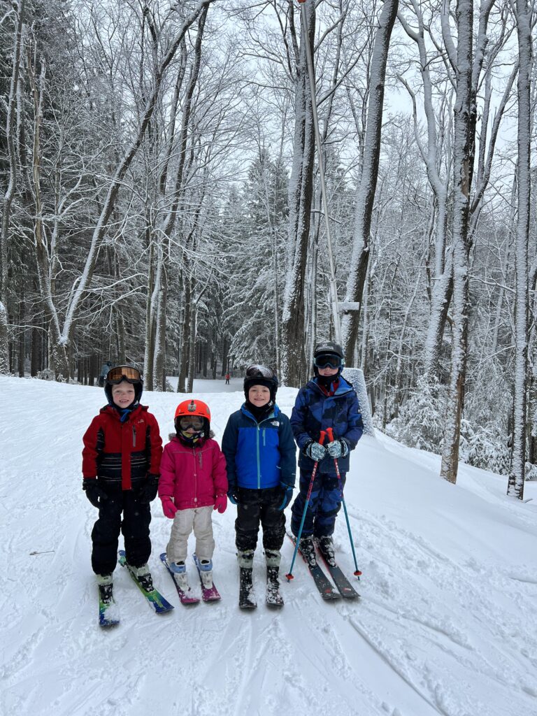 Four children in ski gear — from left: a child in a red jacket, a small child in a pink jacket with an orange helmet, a child in a blue jacket, and an older child in a blue patterned jacket holding ski poles — standing on skis on a snow-covered trail surrounded by tall snow-dusted trees.