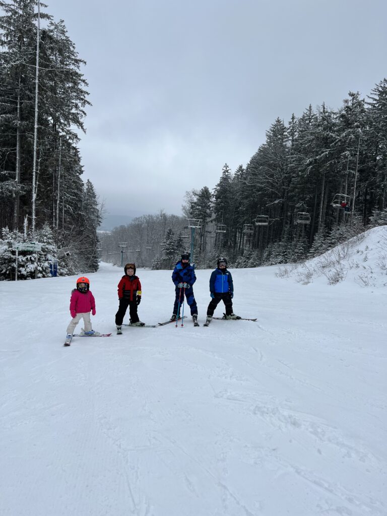 Four children in colorful ski gear standing on a wide snow-covered slope, flanked by tall snow-dusted evergreen trees and empty chairlifts under a gray overcast sky.