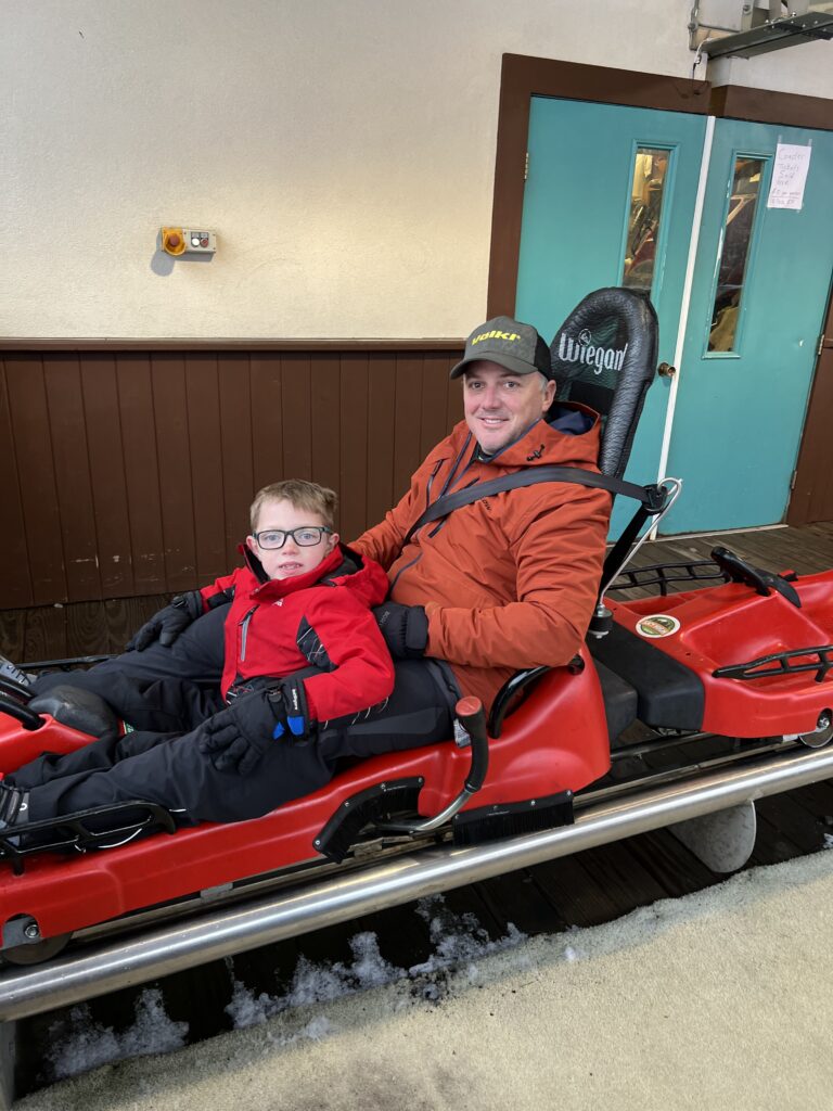 Adult man and young boy sitting together in a red alpine-coaster sled, both wearing winter jackets and gloves — the boy in a bright red jacket and glasses and the man in an orange jacket and black cap, seated in a loading area with teal doors and patches of snow on the platform.