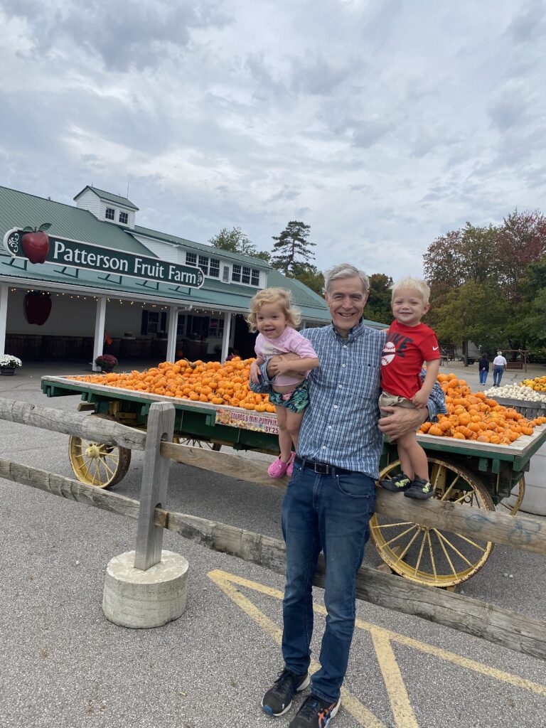 An older man stands by a wooden fence in front of Patterson Fruit Farm, smiling while holding two young children—one in pink and one in red—next to a wagon piled with small orange pumpkins under a cloudy sky.