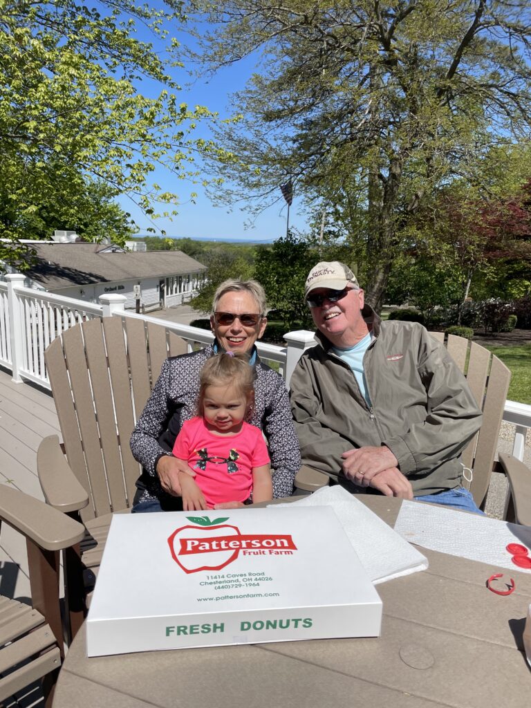 An older couple wearing sunglasses sit on a sunny outdoor patio with a young girl on the woman’s lap, a large Patterson Fruit Farm “Fresh Donuts” box on the table in front of them and trees, white railing and a building visible behind them.