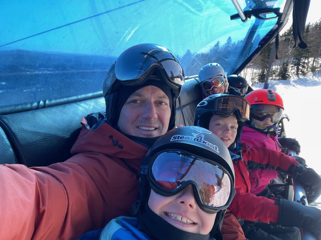 Five people in ski helmets and goggles sit closely together on a covered chairlift, an adult in a red jacket taking a smiling selfie with children in colorful helmets and snowsuits and a snowy slope and evergreen trees visible through the lift's blue canopy.