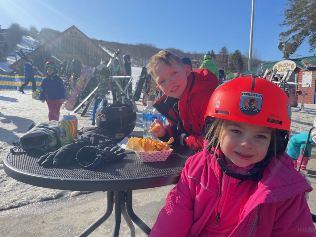 Two children in bright winter clothes sit at a round outdoor table at a snowy ski resort — a smiling girl in a red “Park City, UT” helmet and pink jacket in the foreground and an older child in a red coat reaching for fries from a paper tray, with helmets, gloves, ski racks and a wooden lodge under a clear blue sky in the background.