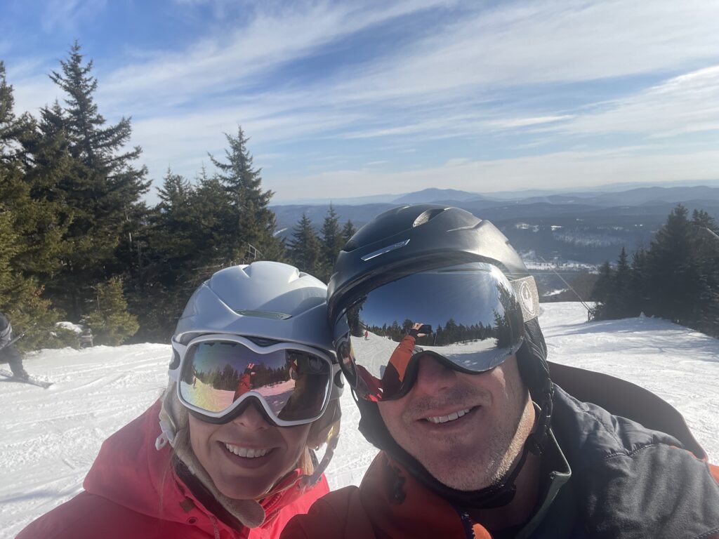 Two smiling skiers in helmets and mirrored goggles take a close-up selfie on a sunlit snowy slope with evergreen trees and distant mountains under a blue sky, the goggles reflecting the slope and the person holding the phone.