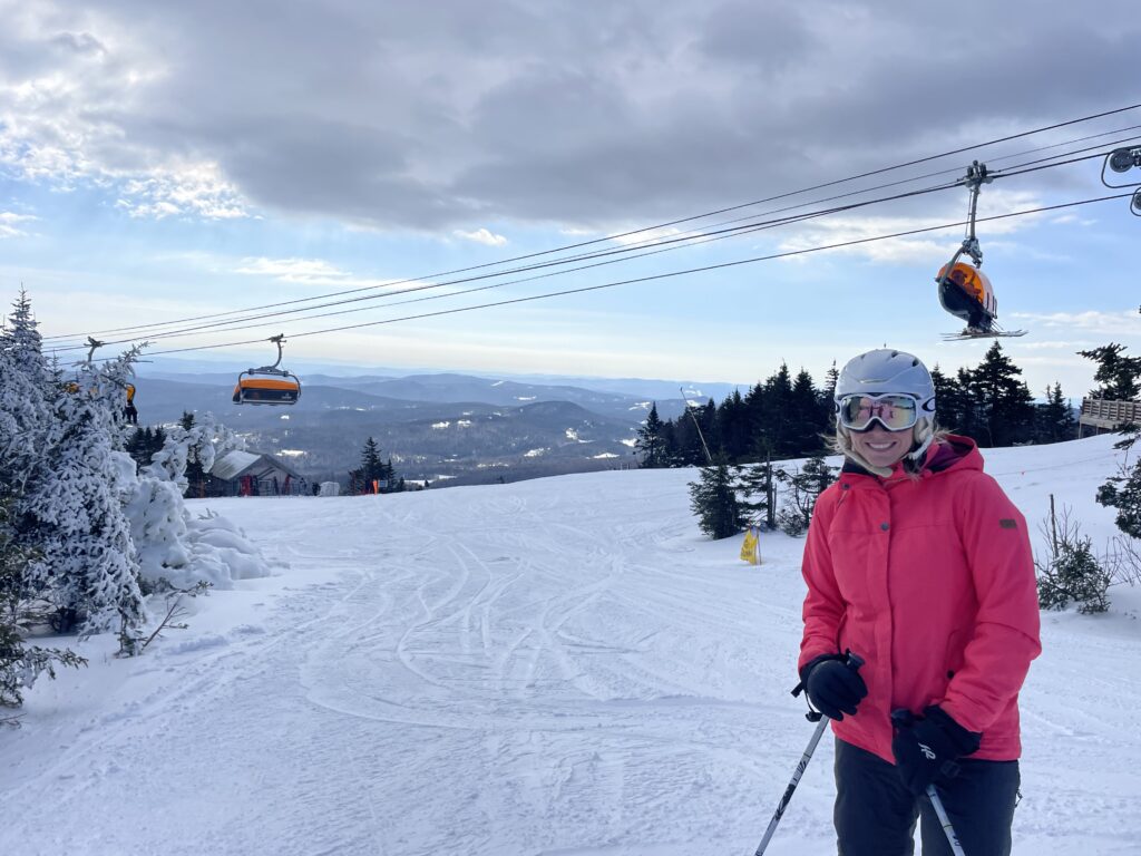 Smiling skier in a bright pink jacket, white helmet and reflective goggles holding poles in the right foreground on a snowy slope with orange chairlift cabins passing overhead, snow-covered trees and distant blue mountain ridges under a partly cloudy sky.