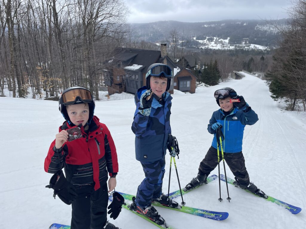 Three boys in helmets and ski gear (red jacket, blue camo jacket, and bright blue jacket) stand on a snowy slope with skis and poles, holding snacks and smiling, with leafless trees, a few mountain homes, and snow-covered hills in the background.