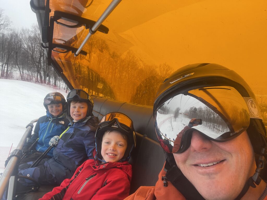 An adult wearing a helmet with mirrored goggles and three smiling boys in ski jackets and helmets sit together on a chairlift under an orange canopy, the goggles reflecting the snowy slope and the photographer's phone with trees visible outside.