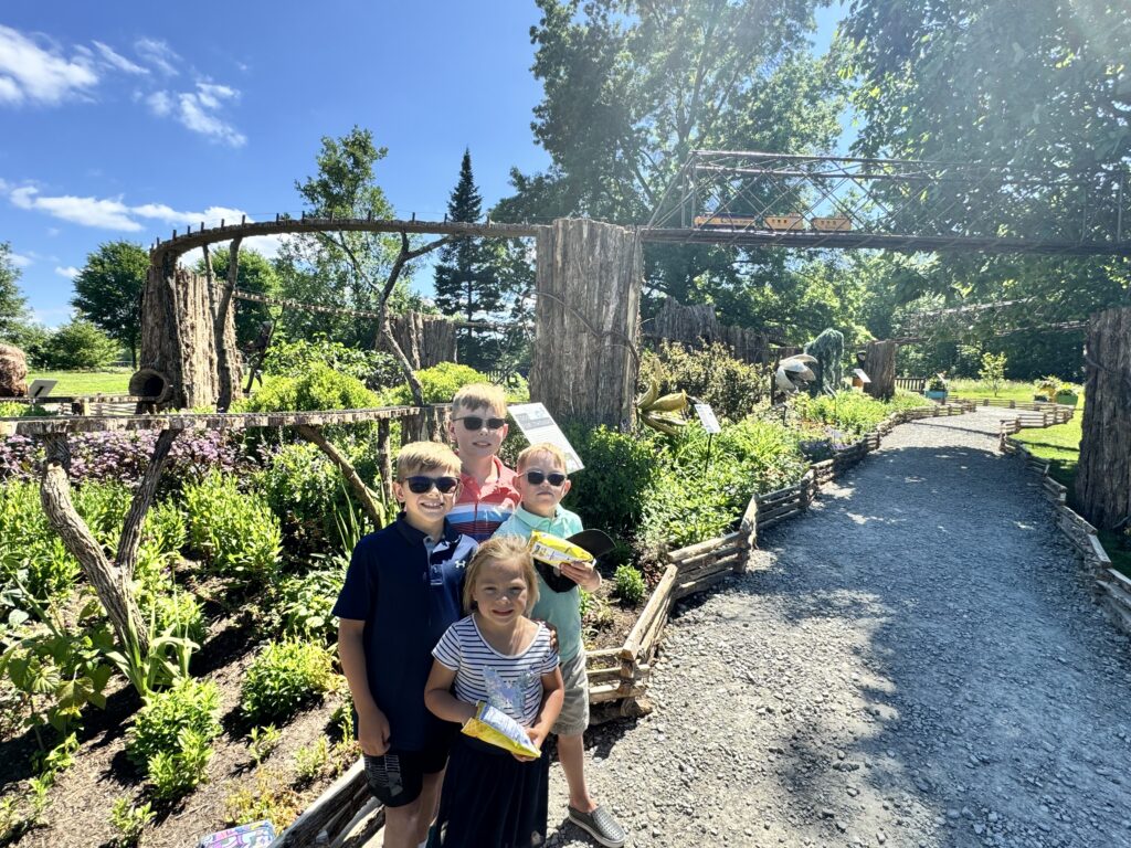 Four smiling children — three boys and one girl — stand together holding snack bags in a sunny botanical garden with raised wooden train tracks, tree-stump supports, and a gravel path winding past flower beds.