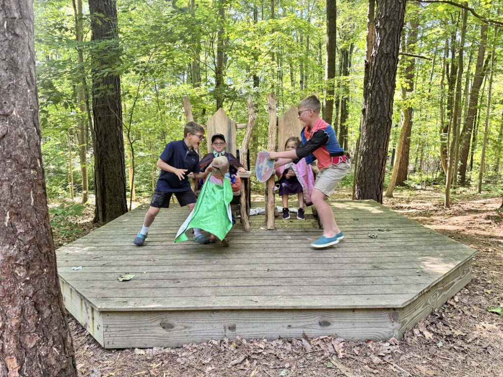 Five children role-playing on a raised octagonal wooden platform in a sunlit forest: one child sits on a rustic wooden throne wearing a cape, another kneels in a bright green cape before them, two boys animatedly act out a confrontation with a painted shield, and a small girl in a pink cape sits beside the throne.