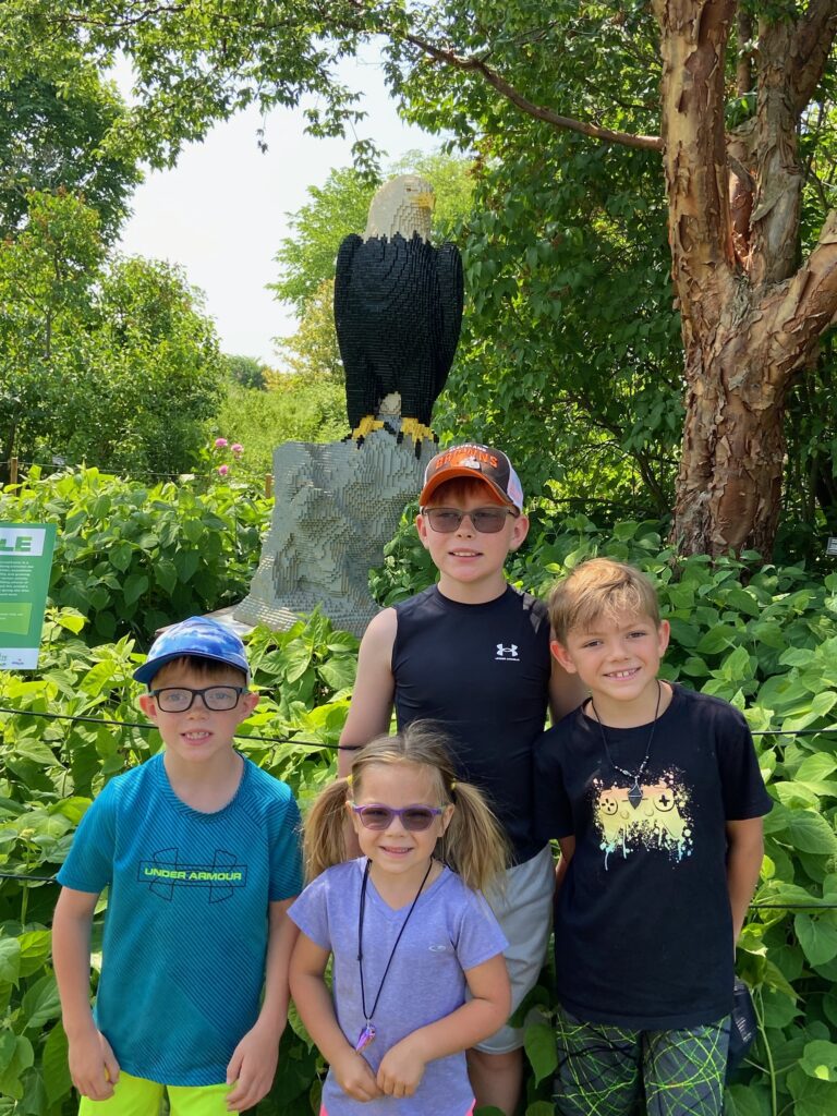Four smiling children — three boys and a girl in a purple shirt with pigtails — standing in front of a large LEGO sculpture of a bald eagle perched on a rock amid lush green foliage.