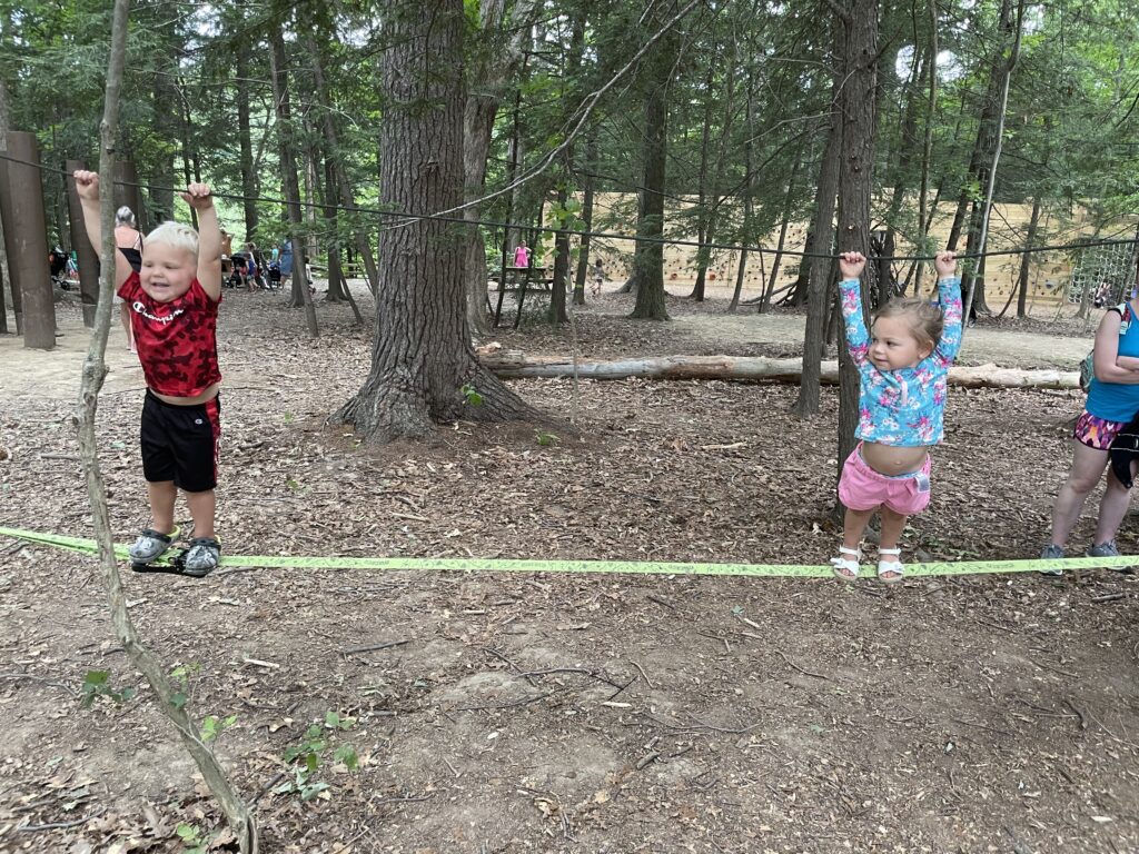 Two young children balancing on a low green slackline between trees in a wooded ropes-course area, each holding an overhead cable—left child in a red patterned shirt and black shorts, right child in a blue floral long-sleeve top and pink shorts, with other people and trees in the background.