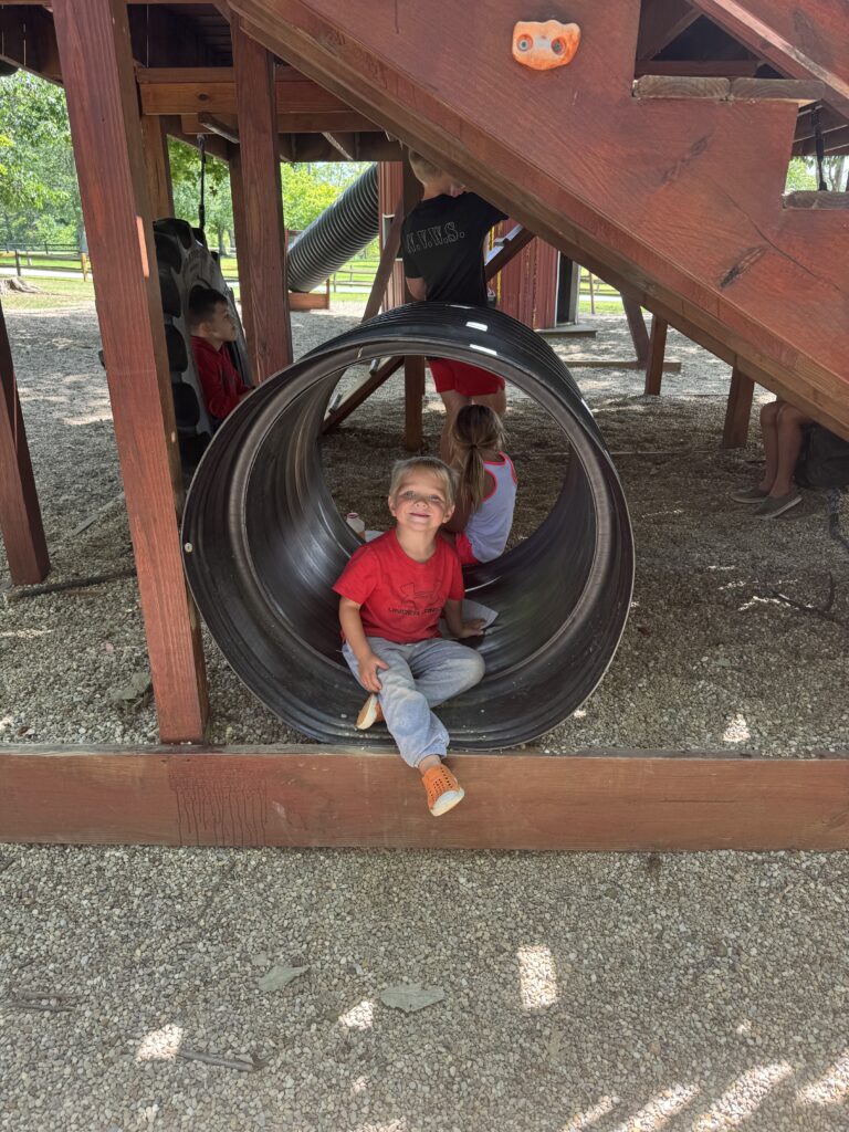 Young boy in a red shirt and orange shoes smiles while sitting in a large black play tunnel under a wooden playground structure, with another child facing away inside the tunnel and other children nearby on gravel.