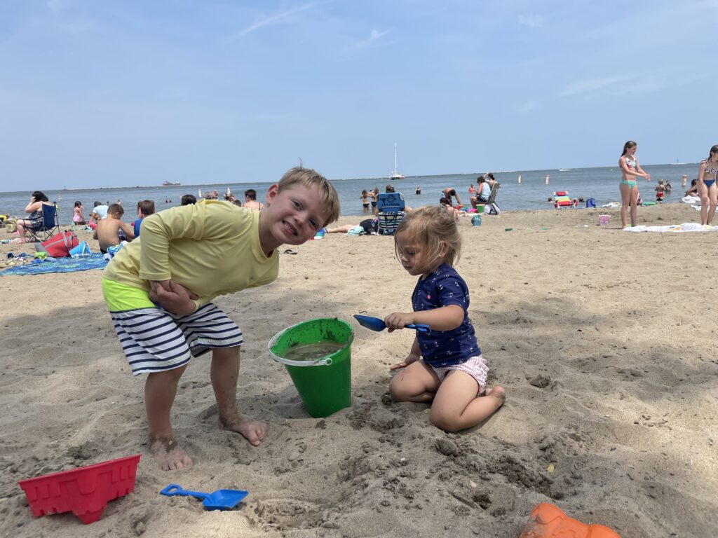 Two young children at a sandy beach — a smiling blond boy in a yellow rash guard and striped swim trunks leaning toward the camera while a toddler girl in a navy shirt and pink ruffled shorts kneels and scoops sand with a blue shovel into a green bucket, with other beachgoers and the ocean visible in the background.