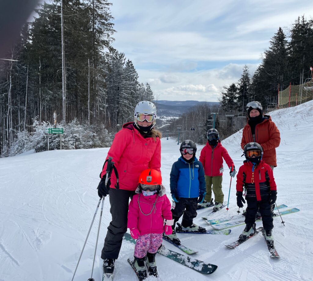 Group of six skiers — two adults and four children — standing and posing on a snowy slope, with the woman in a bright pink jacket and white helmet at left and a man in an orange jacket at right, children wearing colorful jackets and helmets, evergreen trees and a chairlift behind them, and distant snow-covered hills under a cloudy blue sky.