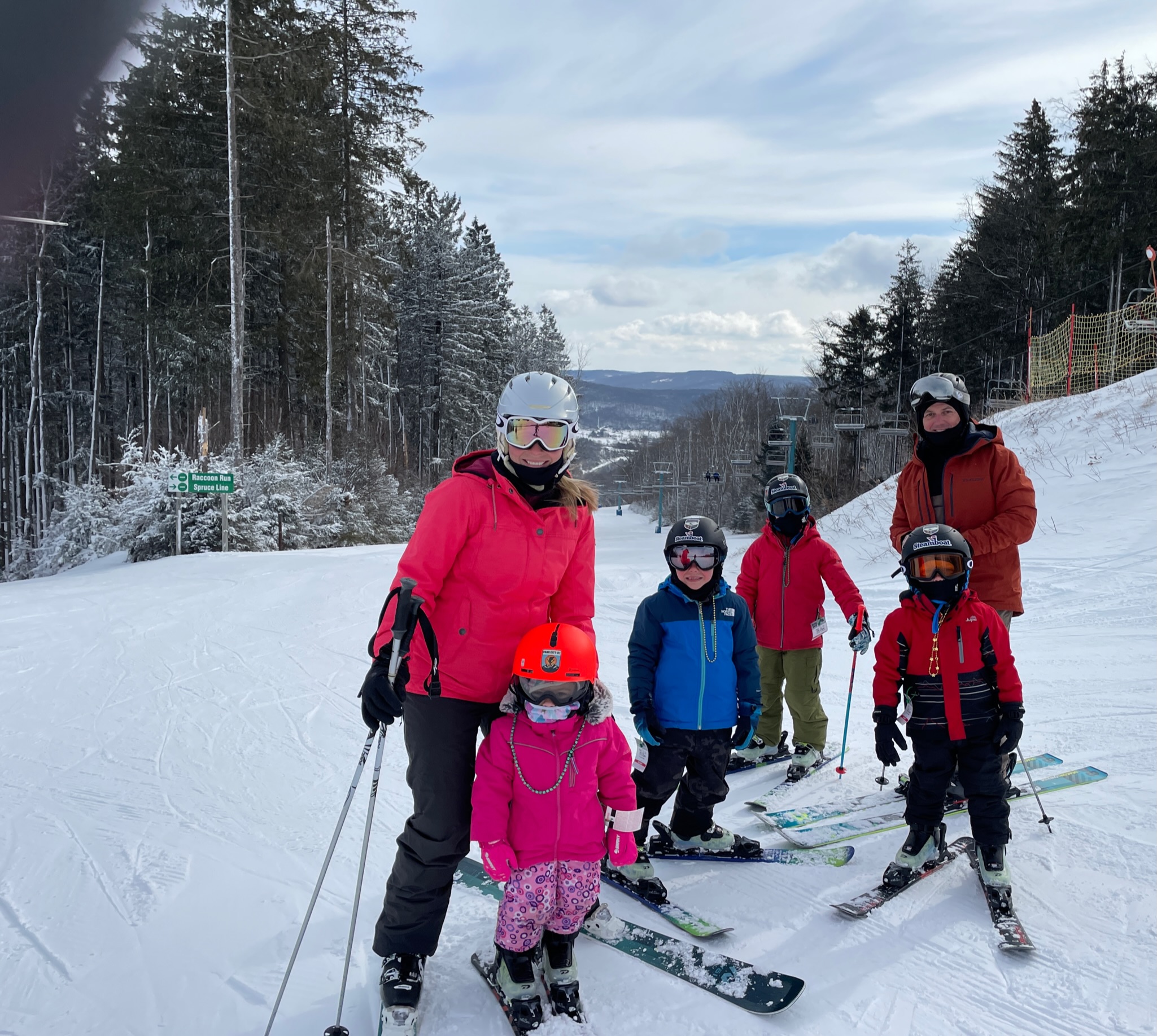 Group of six skiers — two adults and four children — standing and posing on a snowy slope, with the woman in a bright pink jacket and white helmet at left and a man in an orange jacket at right, children wearing colorful jackets and helmets, evergreen trees and a chairlift behind them, and distant snow-covered hills under a cloudy blue sky.