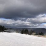 Snow-covered ski run with a snowmaking machine blowing mist near a small group of pine trees, fenced lift buildings and equipment to the right, and heavy dark clouds over distant wooded hills.