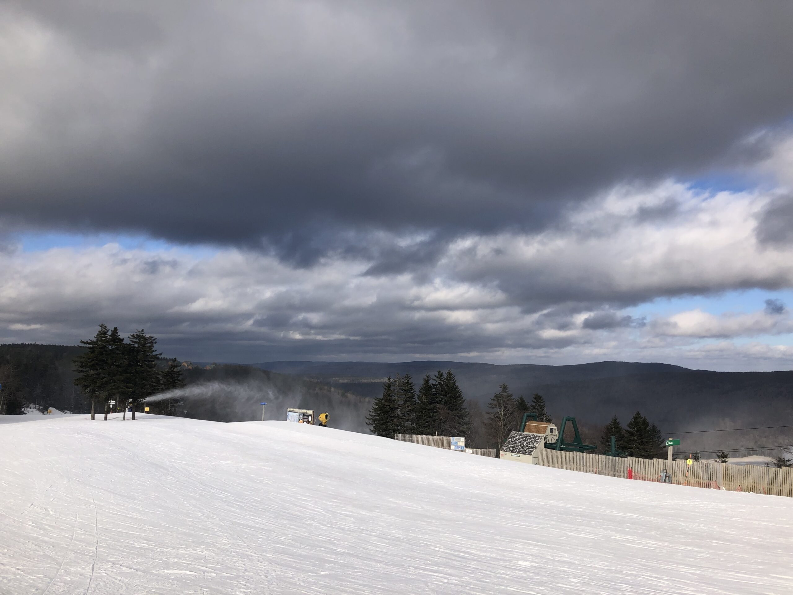 Snow-covered ski run with a snowmaking machine blowing mist near a small group of pine trees, fenced lift buildings and equipment to the right, and heavy dark clouds over distant wooded hills.