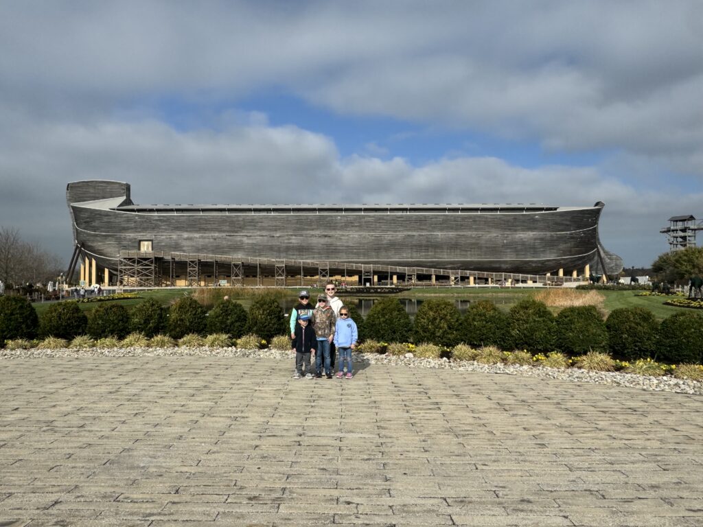 Large timber ark-shaped building on stilts spans the middle of the image under a cloudy blue-gray sky, with a group of five people standing together centered on a wide paved plaza in the foreground and low shrubs, rocks and a small reflecting pond between them.