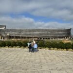 Large timber ark-shaped building on stilts spans the middle of the image under a cloudy blue-gray sky, with a group of five people standing together centered on a wide paved plaza in the foreground and low shrubs, rocks and a small reflecting pond between them.