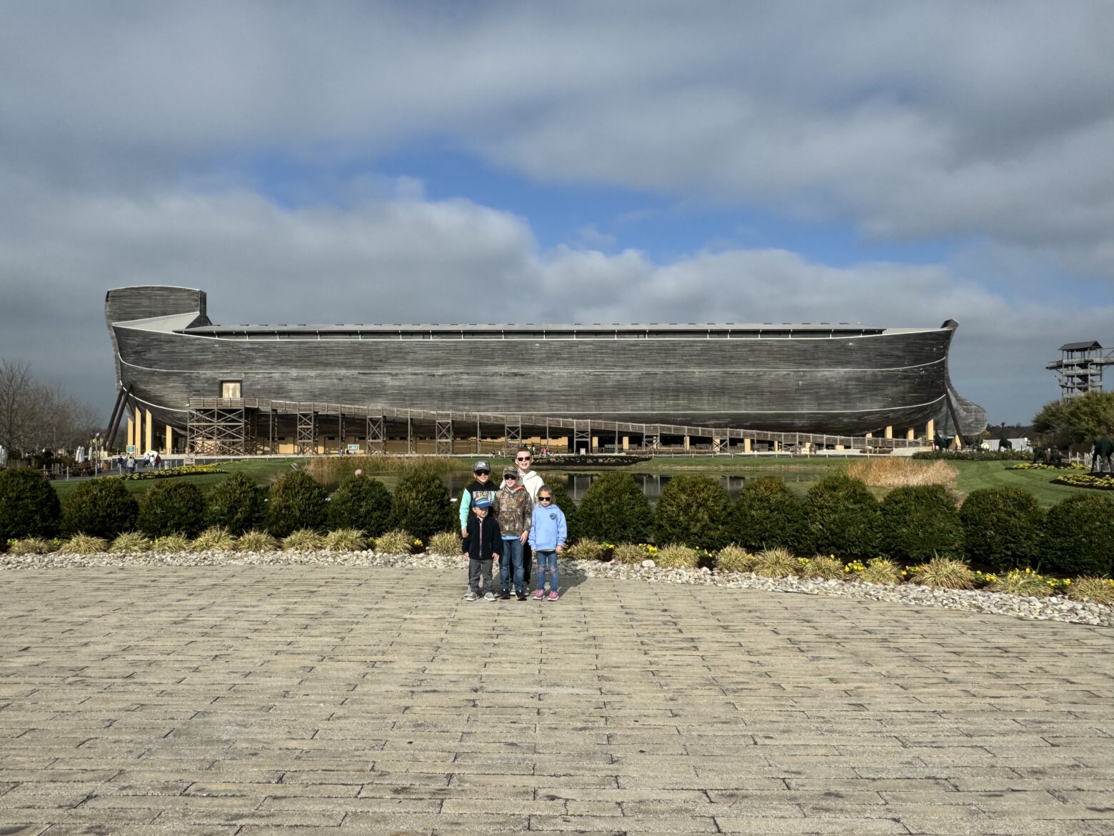 Large timber ark-shaped building on stilts spans the middle of the image under a cloudy blue-gray sky, with a group of five people standing together centered on a wide paved plaza in the foreground and low shrubs, rocks and a small reflecting pond between them.