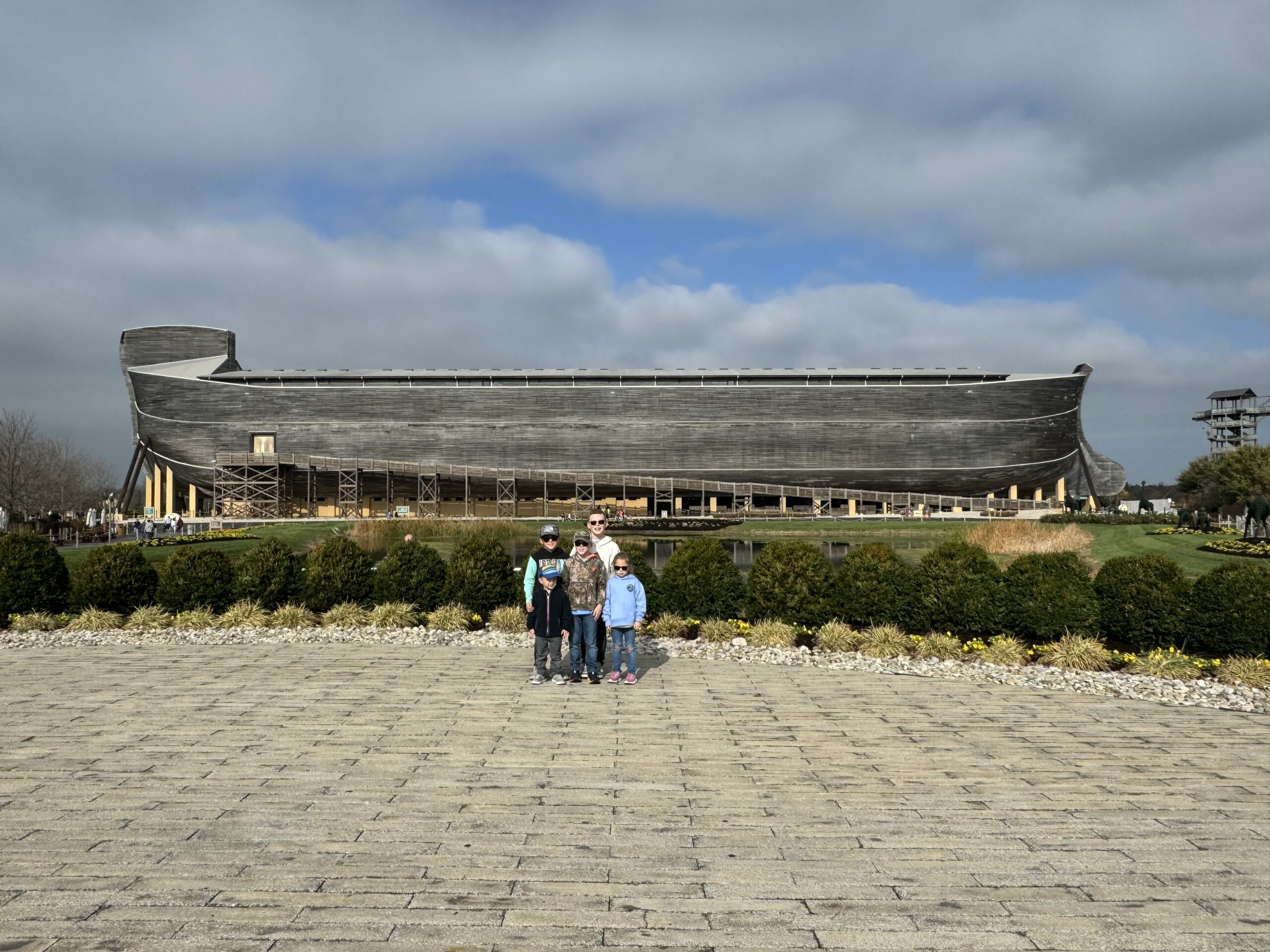 Large timber ark-shaped building on stilts spans the middle of the image under a cloudy blue-gray sky, with a group of five people standing together centered on a wide paved plaza in the foreground and low shrubs, rocks and a small reflecting pond between them.