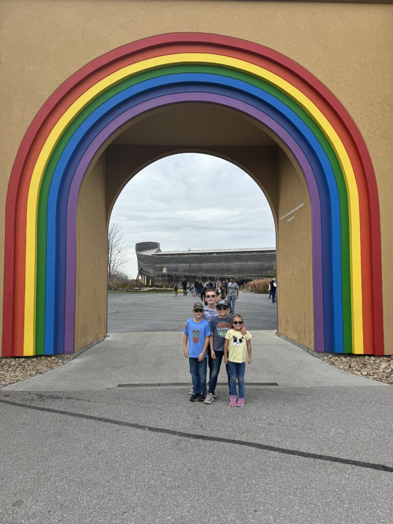 Four children pose beneath a large arch painted in rainbow bands that frames a life‑size wooden ark in the distance with a gray paved plaza and overcast sky.