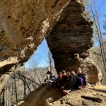 A group of children and a teenager sit on a sunlit sandstone ledge beneath a large, weathered rock arch, with bare trees and a clear blue sky visible in the forest beyond.