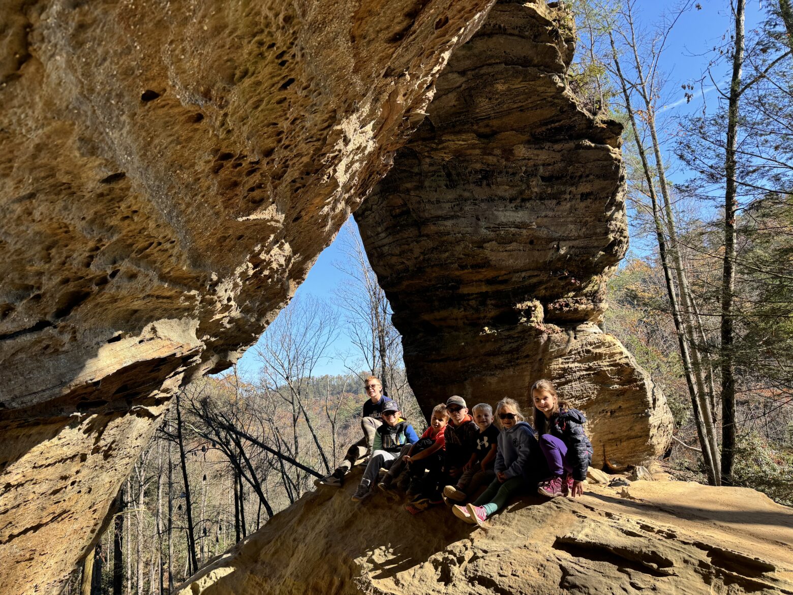 A group of children and a teenager sit on a sunlit sandstone ledge beneath a large, weathered rock arch, with bare trees and a clear blue sky visible in the forest beyond.