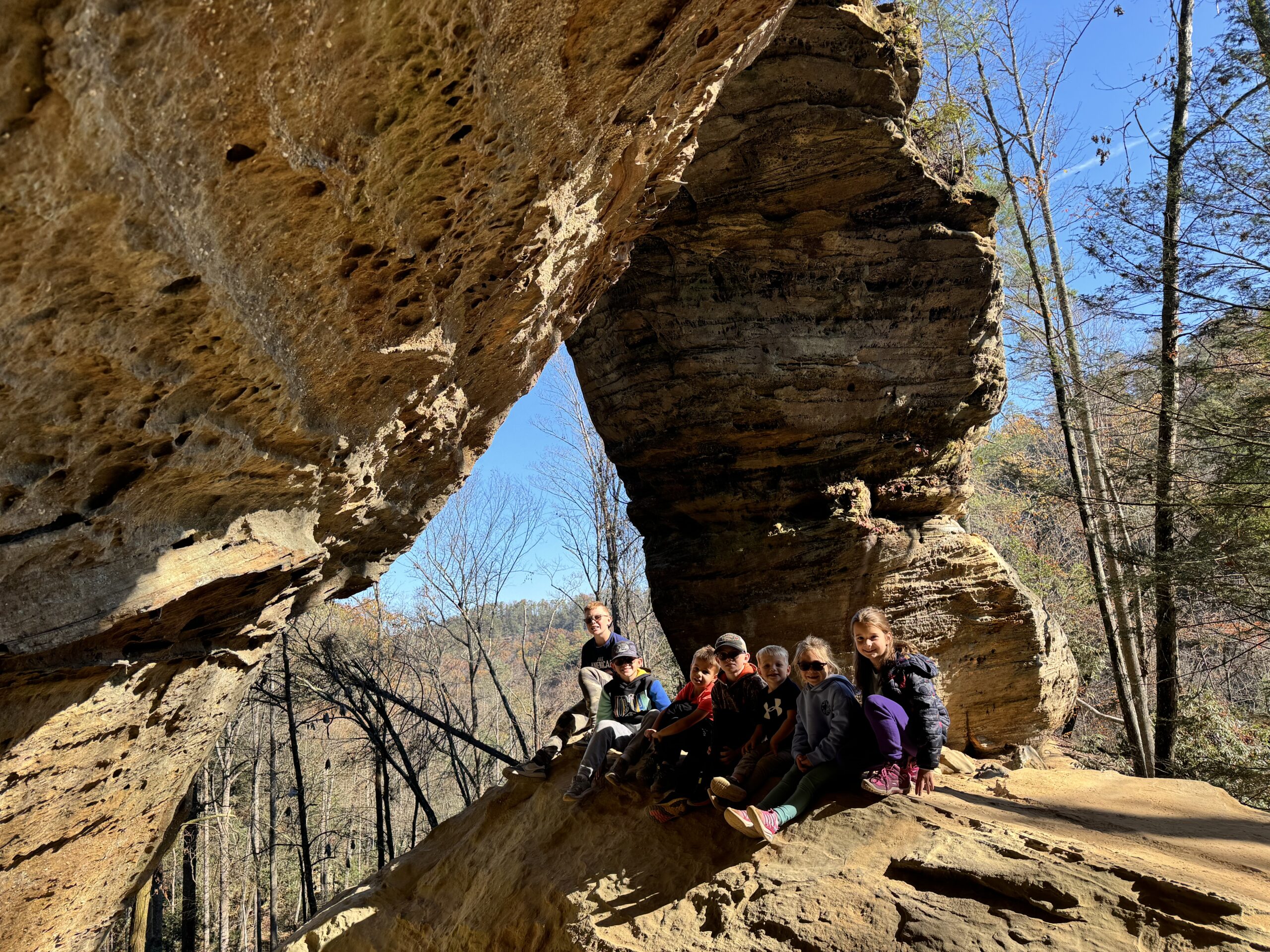 A group of children and a teenager sit on a sunlit sandstone ledge beneath a large, weathered rock arch, with bare trees and a clear blue sky visible in the forest beyond.