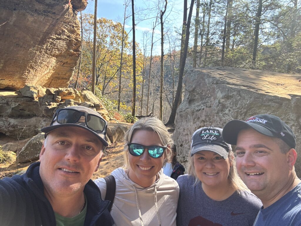 Four smiling adults — two men and two women — take a selfie in a sunlit forest clearing in front of large sandstone rock formations, one woman wearing mirrored teal sunglasses and another wearing a 'Lake Life' baseball cap.