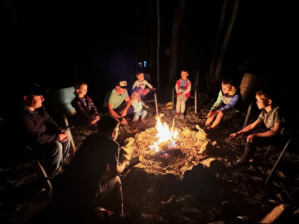 A group of adults and children sit in a circle of chairs around a bright campfire at night in a wooded area, their faces and the rocky fire pit lit by warm orange flames.