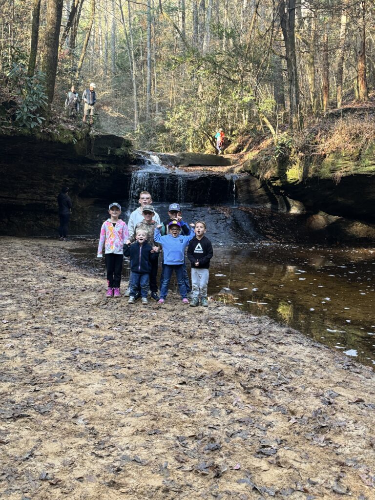 A group of children posing on a leaf-covered sandy bank in front of a small waterfall flowing over a rocky ledge in a wooded area, with a few adults and hikers visible on the rocks above.