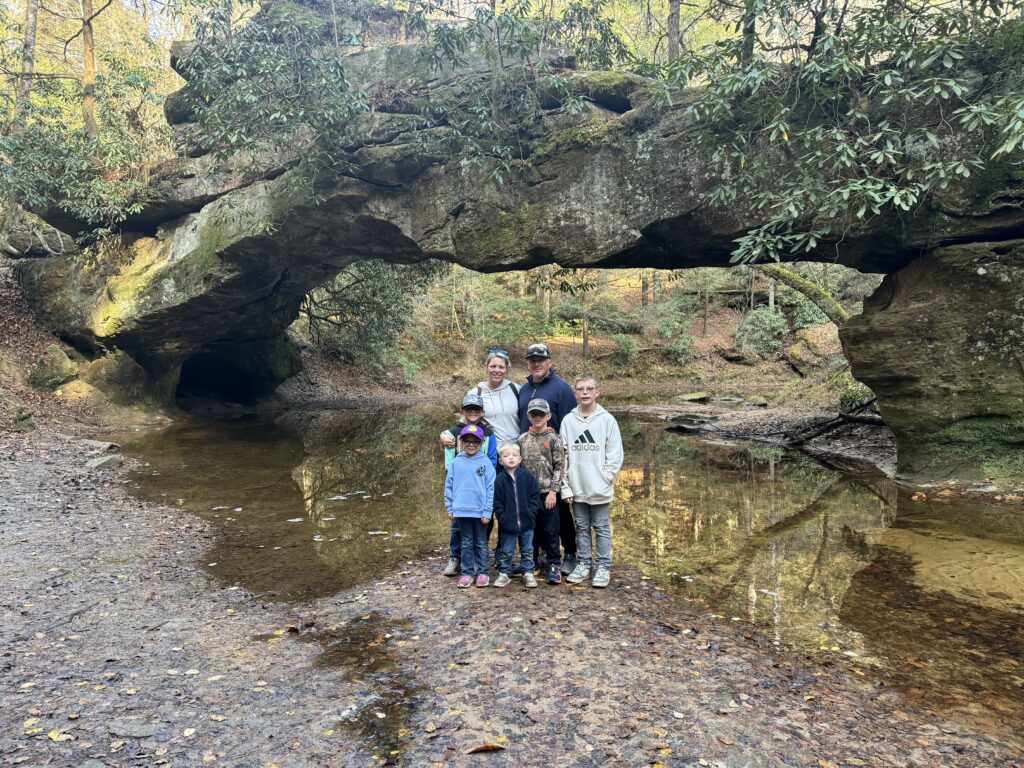A family of seven poses on a muddy riverbank beneath a large mossy sandstone natural arch spanning a shallow stream, with trees and autumn foliage reflected in the water.