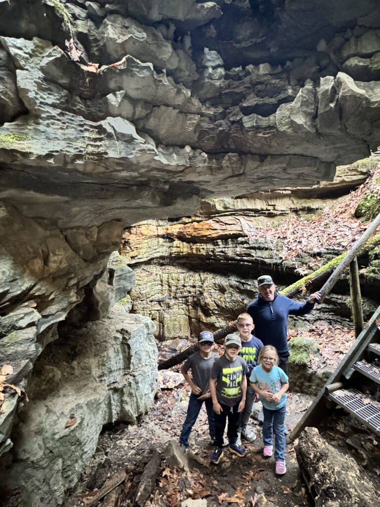 An adult man and four children stand together under a large layered sandstone overhang in a forested rock shelter, on leaf-strewn ground next to a metal stairway.