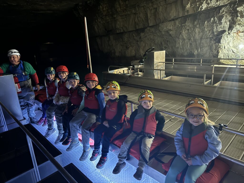 Seven children wearing helmets and red life jackets sit side-by-side on a metal bench inside a dimly lit limestone cave or underground boat platform, with an adult crew member at the far left and rocky walls and metal railings illuminated in the background.